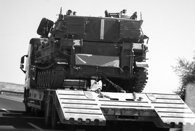 Black and white shot of a heavy military tracked vehicle loaded onto a large flatbed truck trailer on a highway.
