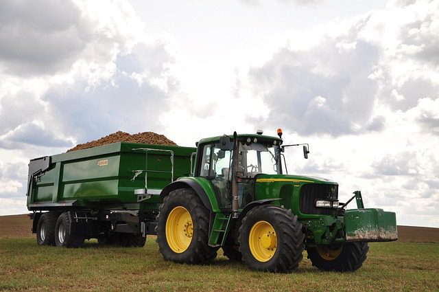 A green John Deere tractor, possibly a 6100 model, with a full trailer attached, parked in a field under a cloudy sky.